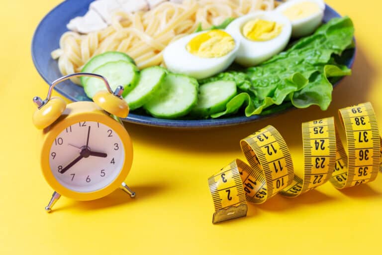 Plate with food and alarm clock on yellow background, interval fasting concept