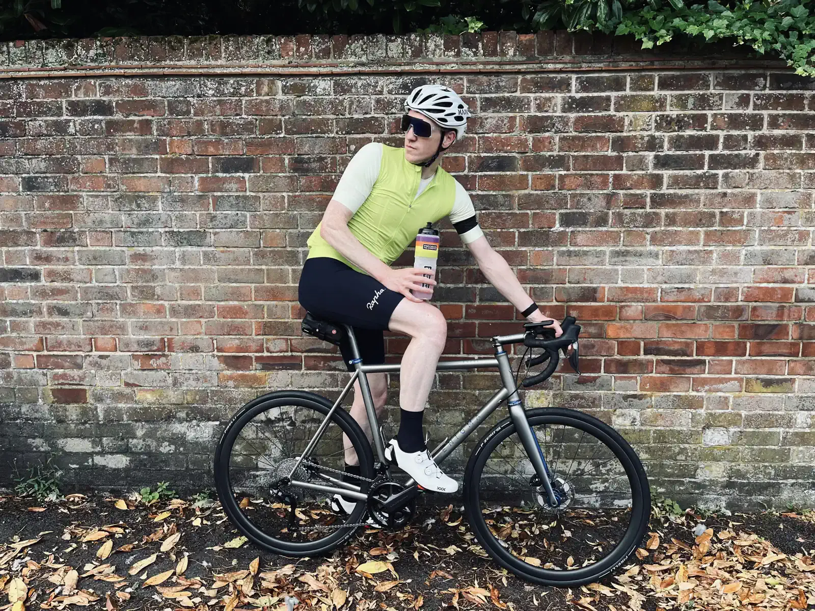 A cyclist in a yellow shirt and helmet sitting on a road bike by a brick wall, holding a water bottle.