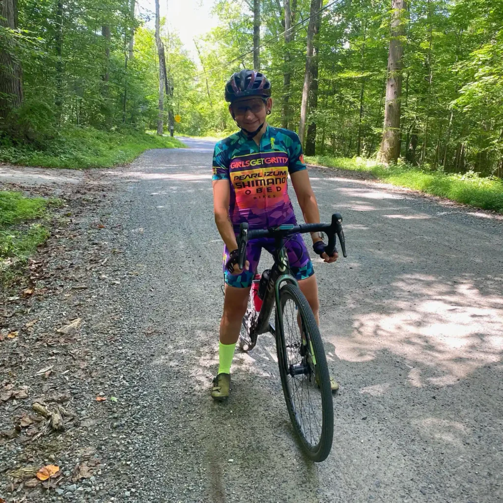 A cyclist in colorful attire standing with a bicycle on a shady forest road.