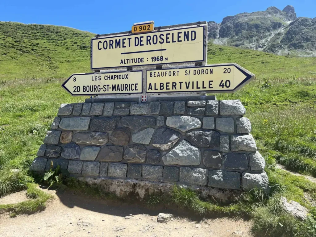 Directional signpost at cormet de roselend indicating altitude and distances to nearby localities against a backdrop of mountains and blue sky.