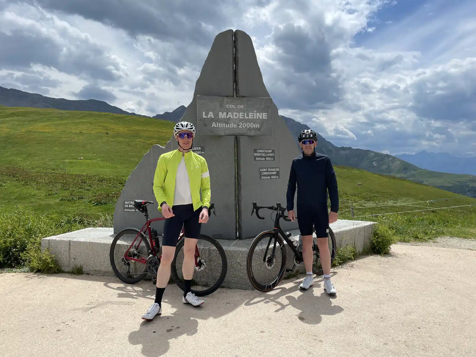 Two cyclists standing by a monument at col de la madeleine, at an altitude of 2000m, with scenic mountain views in the background.