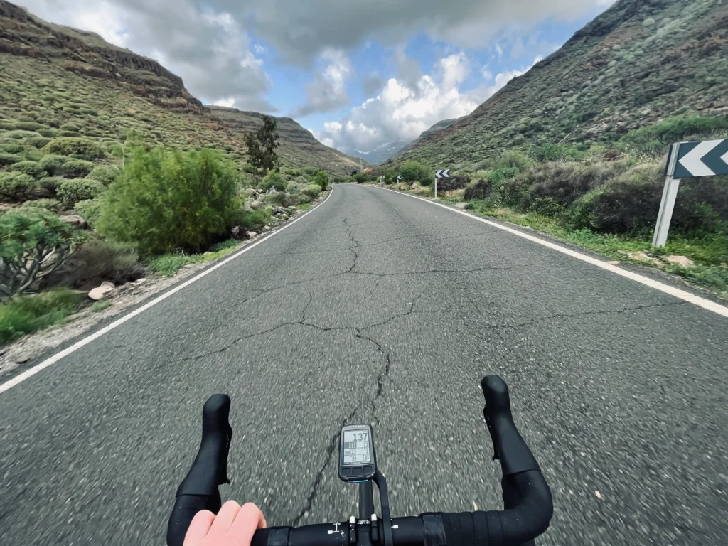 First-person view from a bicycle on a mountain road with cracked asphalt, surrounded by lush hills under a cloudy sky, featuring a handlebar with a mounted bike computer.