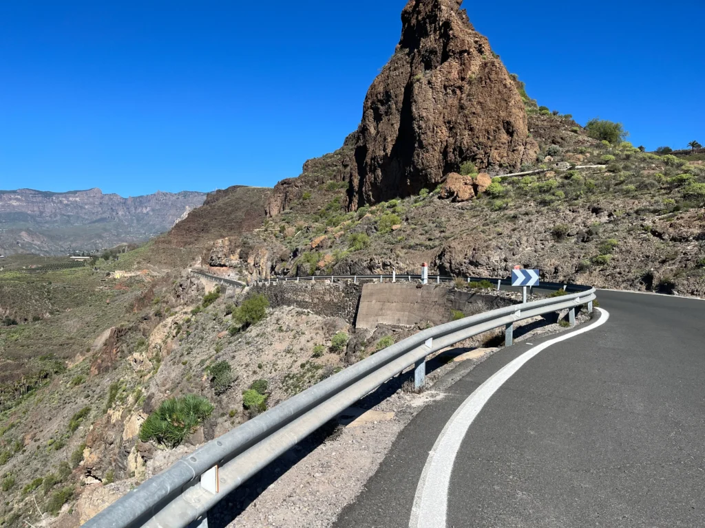 Winding road with guardrails through mountainous terrain, featuring a large rocky outcrop on the right and clear blue sky.