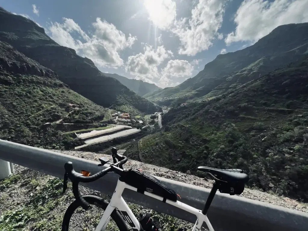 A road bike leaning on a guardrail overlooking a sunlit mountainous landscape with a small village nestled in a valley.