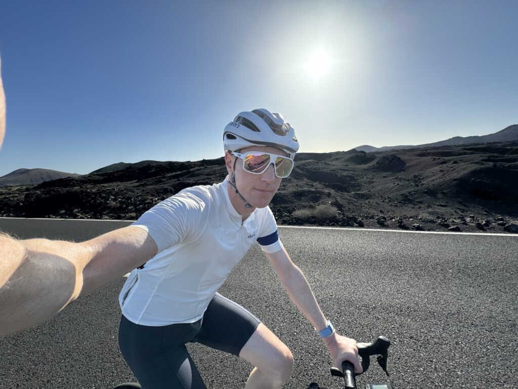 A person wearing cycling gear and a helmet is riding a bike on a paved road with a rocky landscape in the background, under a clear, sunny sky.