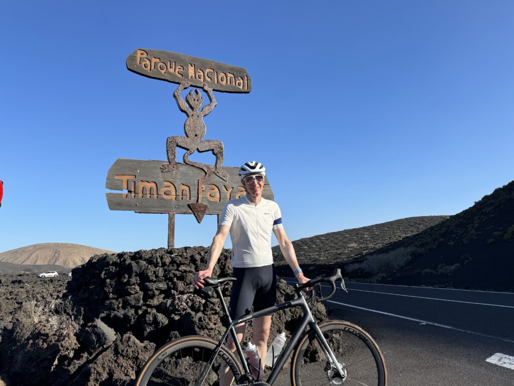 A cyclist stands with a bike in front of a wooden sign for Parque Nacional Timanfaya on a clear day. The background features a rocky landscape and a clear blue sky.