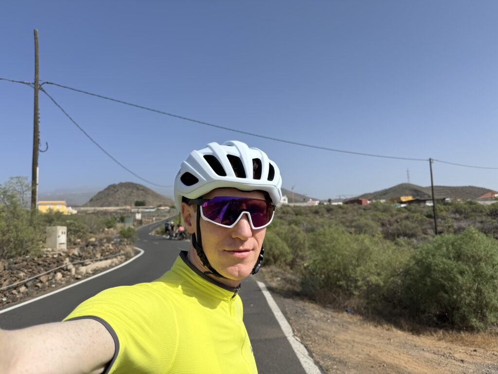 A cyclist wearing a white helmet, yellow shirt, and sunglasses takes a selfie on a rural road with hills and houses in the background. The sky is clear and sunny.