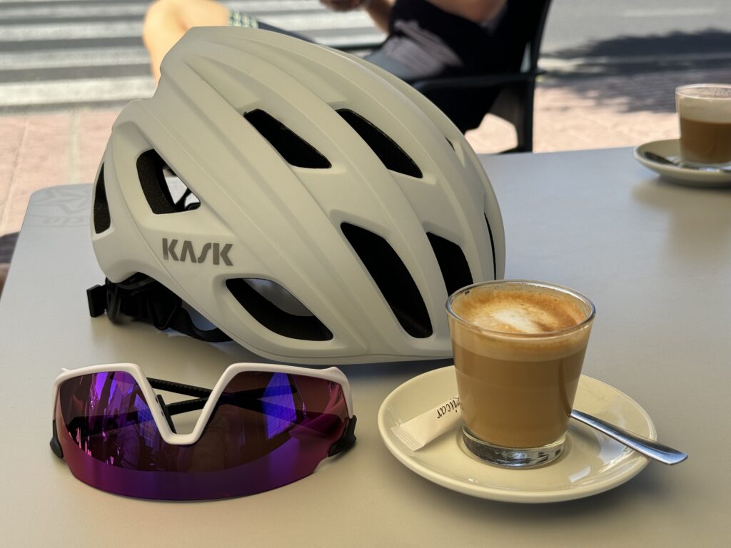 A white cycling helmet, a pair of purple-tinted cycling glasses, and a cup of cappuccino on a saucer with a spoon are placed on a table outdoors.