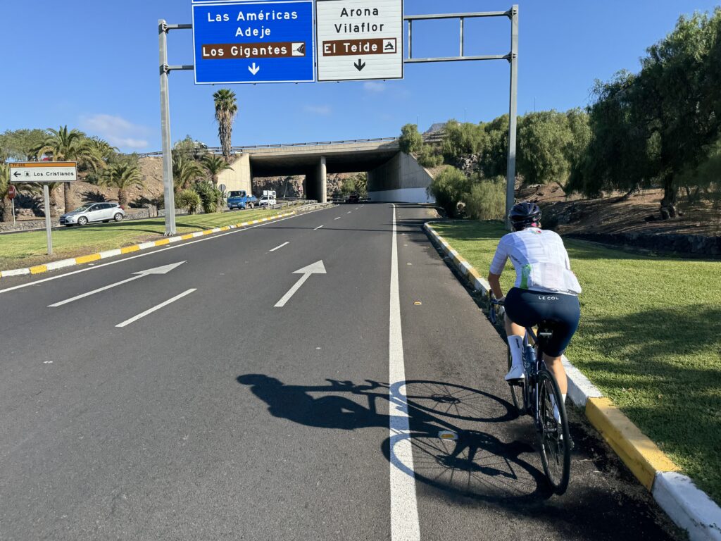 A cyclist rides on a paved road towards an overpass, with signs overhead indicating directions to various destinations including Las Américas, Adeje, Los Gigantes, Arona, Vilaflor, and El Teide.