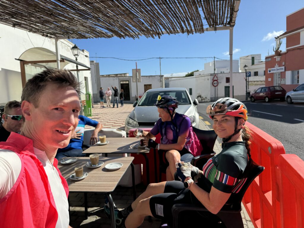 Four cyclists sit at an outdoor café table drinking coffee. A man in a red jacket takes a selfie. The background includes white buildings and a parked car under clear blue skies.