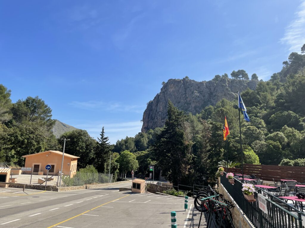 Outdoor scene of a small building and a parking lot with a mountainous landscape in the background. There are several bicycles parked on the right, and flags flying near a patio area.