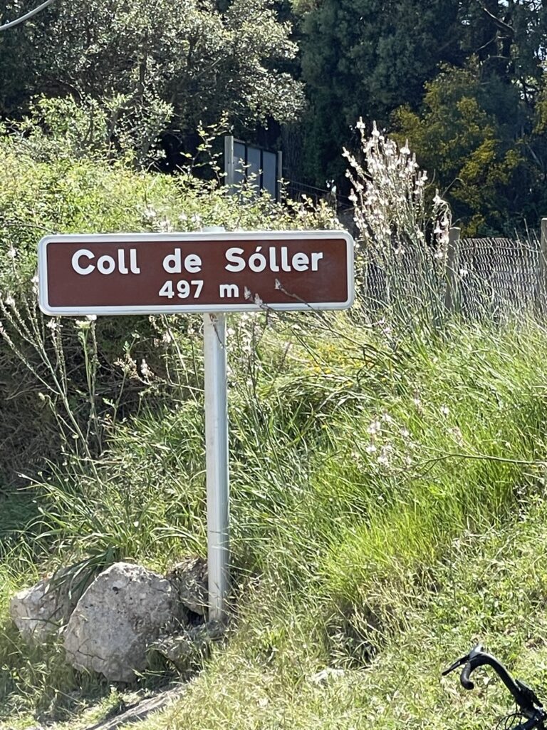 A brown and white sign reads "Coll de Sóller 497 m" placed on a grassy roadside with vegetation and a partial view of a wire fence in the background.