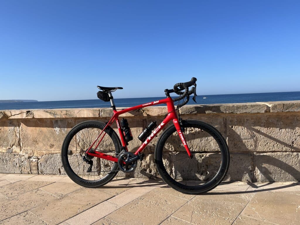 A red road bike is parked against a stone wall by the seaside on a sunny day, with a clear blue sky and ocean in the background.