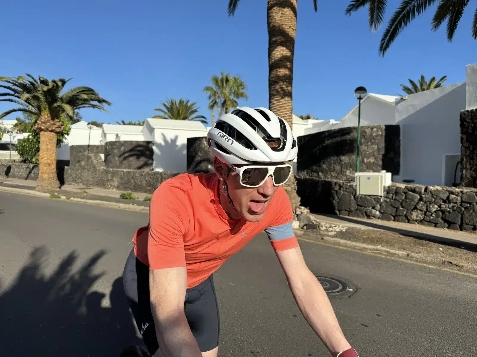 A cyclist in an orange shirt, white helmet, and sunglasses rides on a sunny street with palm trees and white buildings in the background.