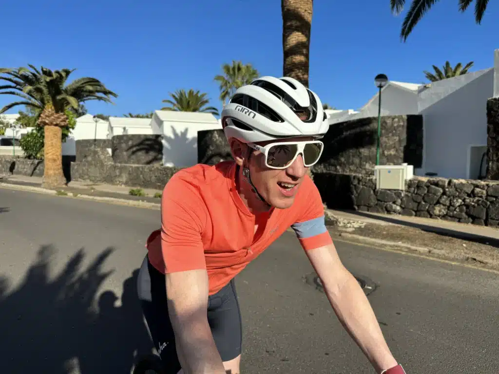 A cyclist in an orange jersey and white helmet rides down a sunny street lined with palm trees and white buildings.