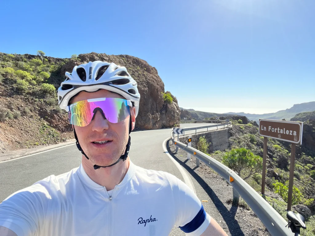 Cyclist in a white jersey and helmet taking a selfie on a sunny road near mountains, with a sign reading "La Fortaleza.