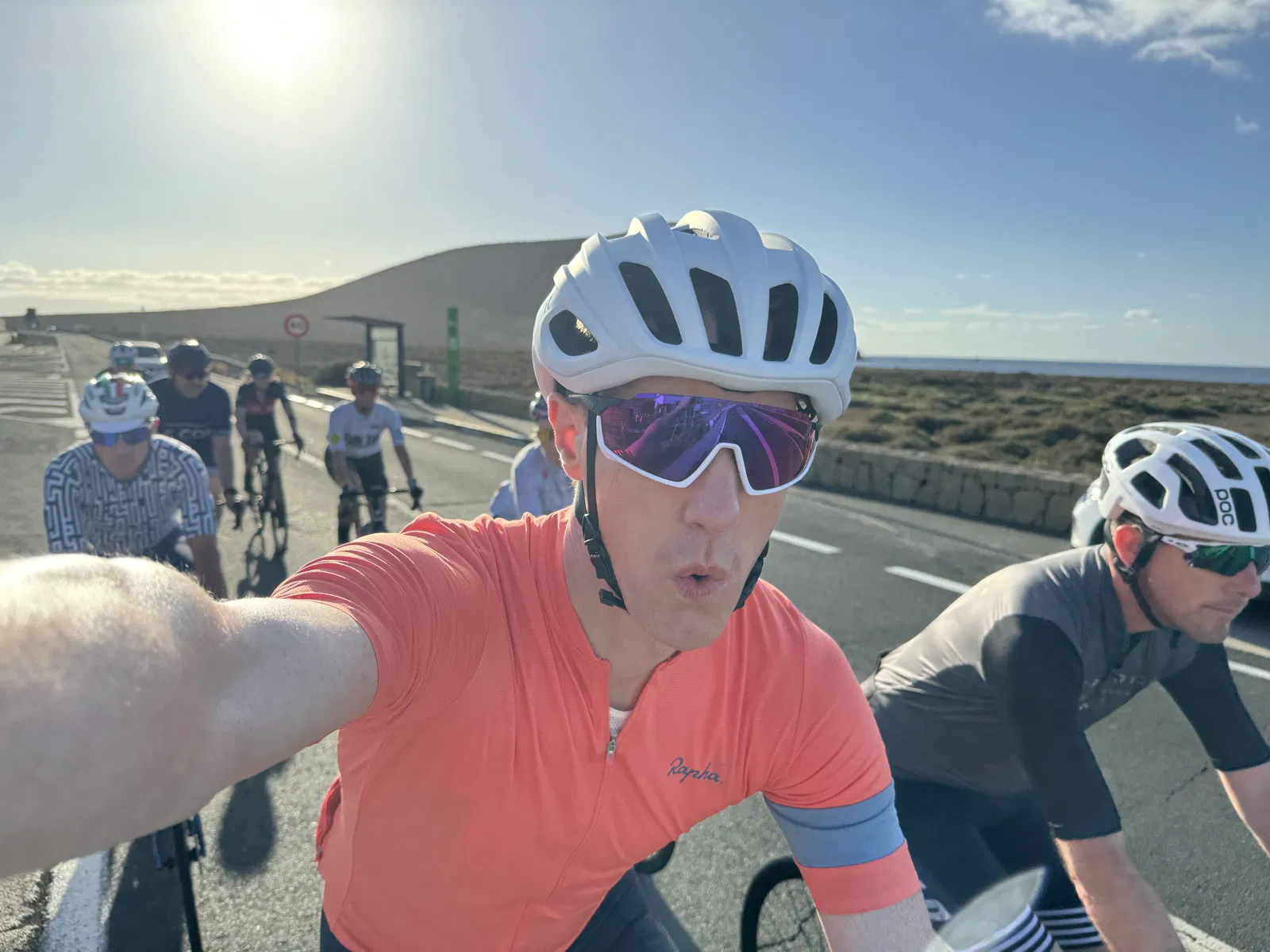 A group of cyclists riding on a paved road with a clear sky in the background. The selfie captures a man in an orange cycling shirt in the foreground with others following behind.