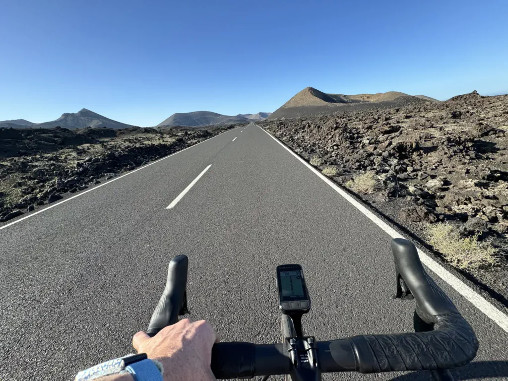 A person rides a bike on an empty, paved road through a rocky, mountainous landscape under a clear blue sky.