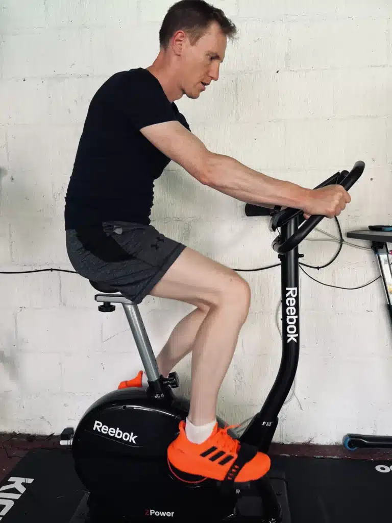 A person in a black shirt and gray shorts rides a Reebok stationary bike indoors. They are wearing bright orange athletic shoes.