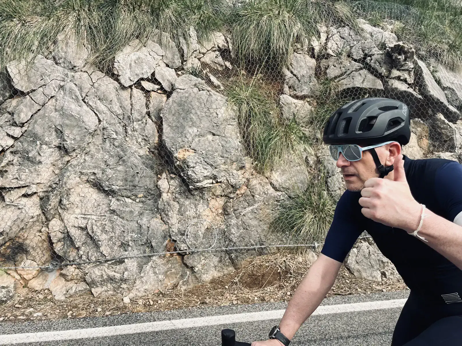 A cyclist in a black helmet and shades gives a thumbs-up while riding by a rocky hillside.