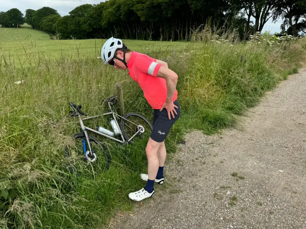 A biker in a red jersey and white helmet is checking out their bike that’s toppled over by the grassy roadside.