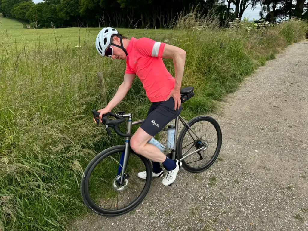 A guy in a red shirt, black shorts, and a helmet is hanging out next to his bike on a country path. He seems to be tweaking or checking something near the back wheel. There’s greenery all around and he’s on a dirt road.