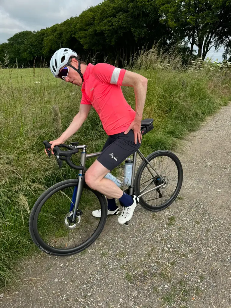 A cyclist rocking a pink jersey and helmet is standing by his bike on a gravel path, grabbing his lower back and slightly leaning forward.