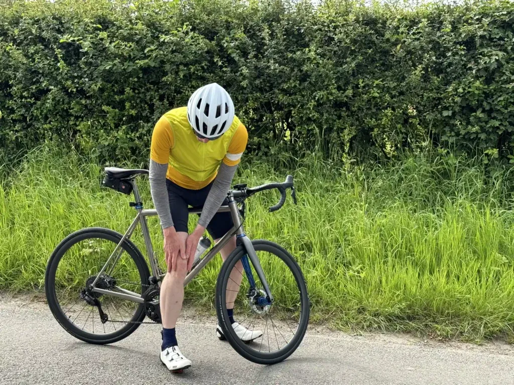 A cyclist in a yellow jersey and white helmet is bent over their bike, clutching their knee, on a road next to some thick bushes.