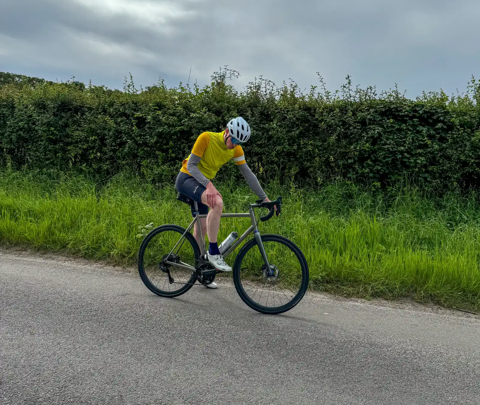 A dude in a yellow jersey and helmet is biking along a paved road with green scenery around, under a cloudy sky.