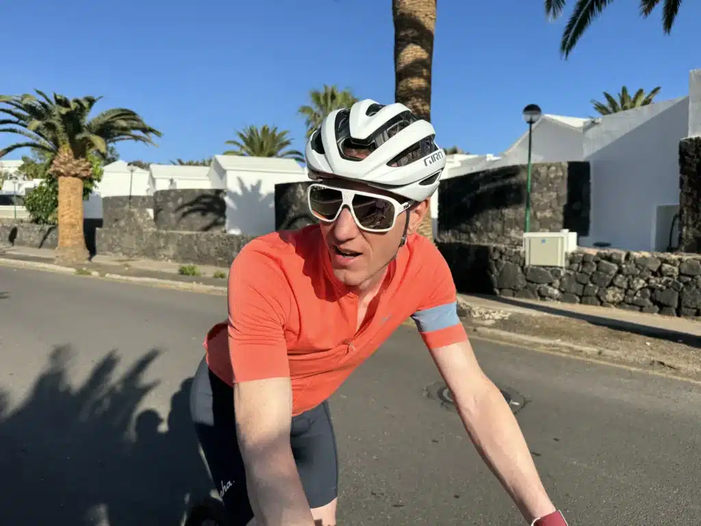 A person in a red cycling shirt and white helmet rides a bicycle on a sunny day with palm trees and white buildings in the background.