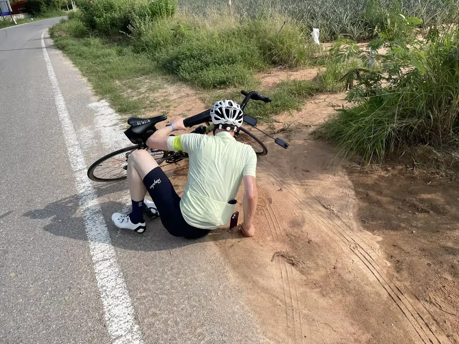 A cyclist in full gear is chilling on the ground by their bike, which looks like it took a tumble. They're just hanging out next to some grass on the roadside.