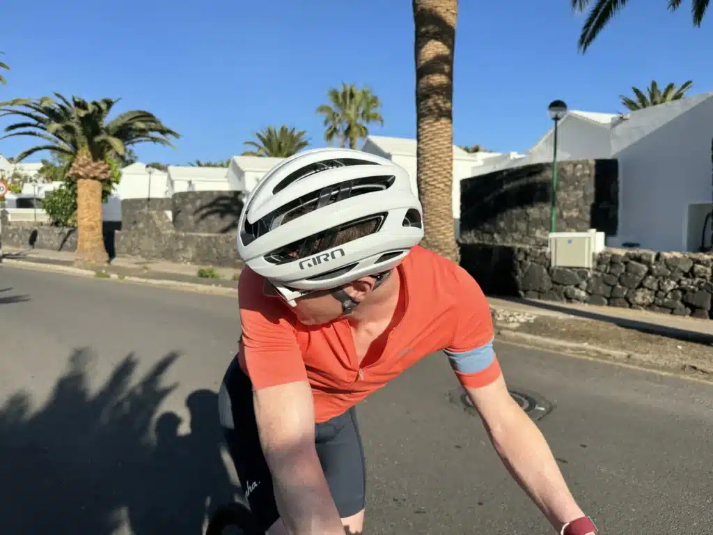 A cyclist in an orange shirt and white helmet looks down while riding on a sunny day, with palm trees and white buildings in the background.