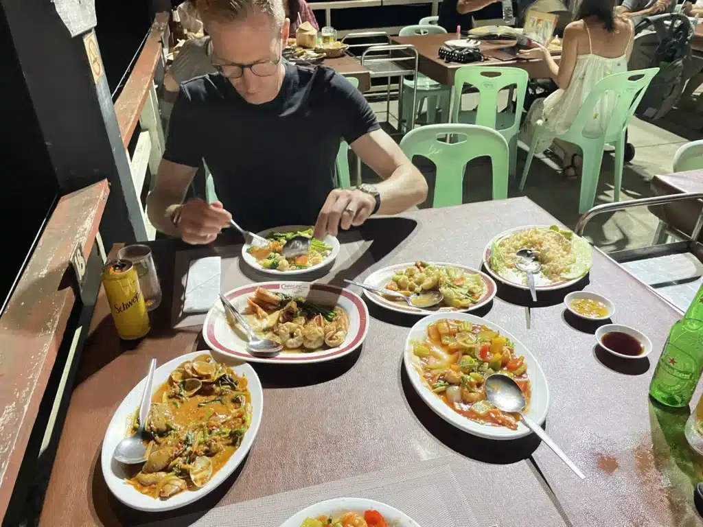 So, there's this person chillin' in a black shirt, munching on a meal with all sorts of goodies like curry and veggies, plus a drink. They're outside at some dining spot with other folks scattered around at nearby tables.