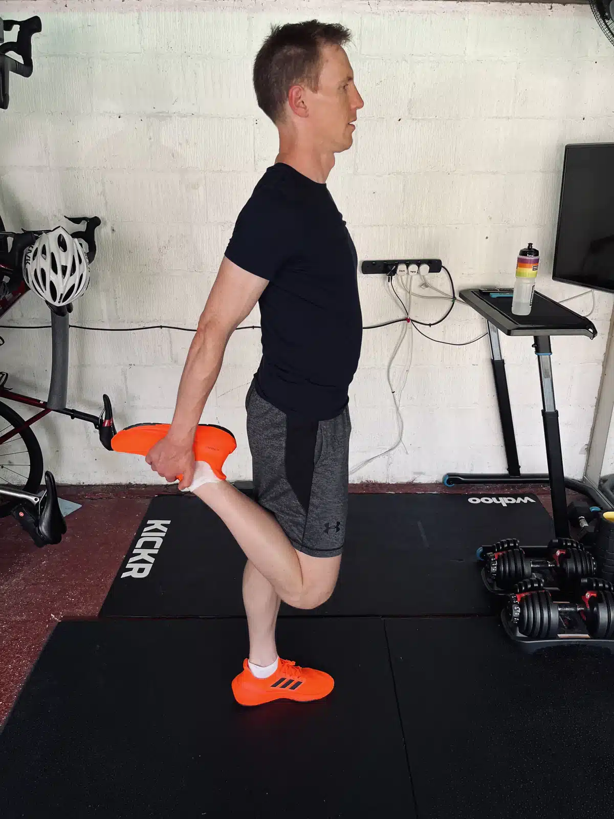 A person in sportswear is standing on one leg, holding their other foot behind them in a quadricep stretch. Exercise equipment like a bike and weights are around them.