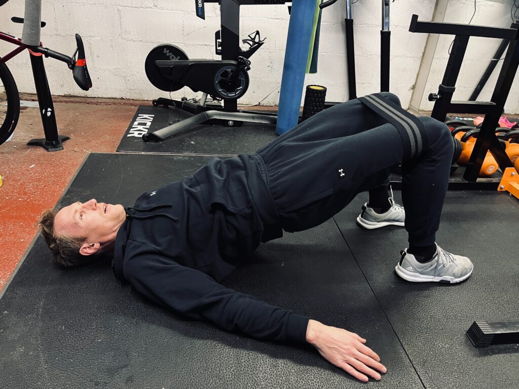 Someone decked out in workout gear is doing glute bridges with a resistance band in their home gym. You can see a bunch of other gym equipment scattered around in the background.