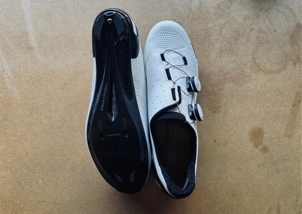 So, there's this pic of some white cycling shoes with black soles. They're lying next to each other on a light brown surface. One shoe's facing down so you can check out the sole, while the other is showing off its top view with two dials for tightening.