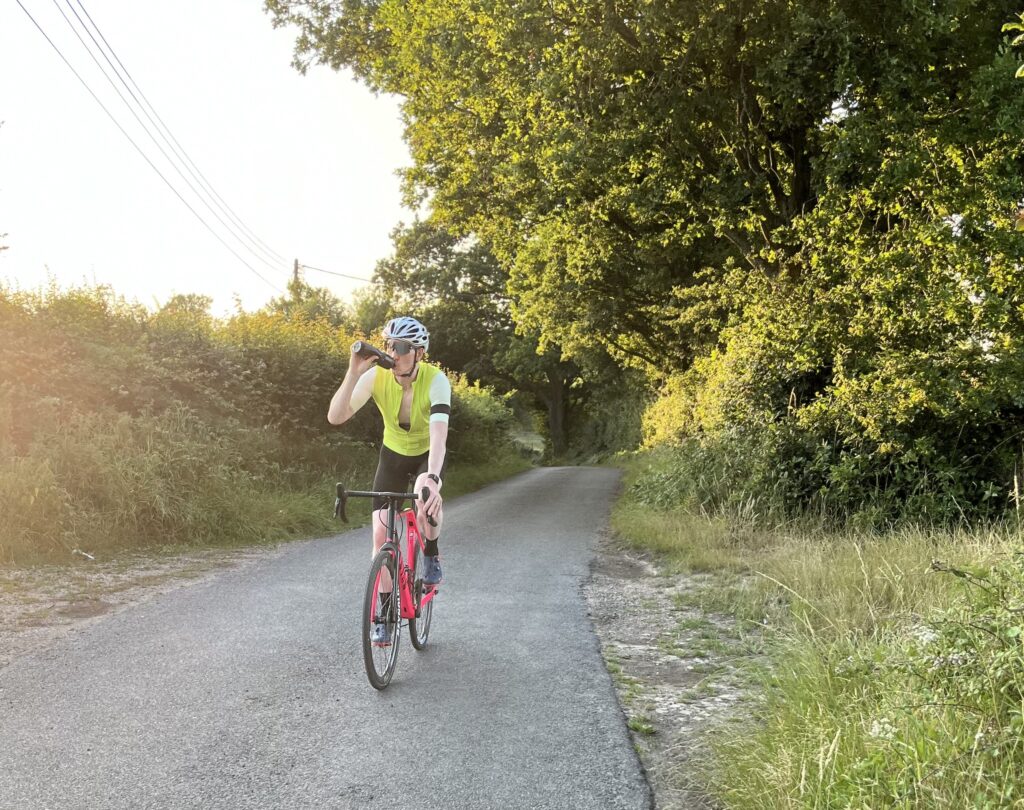 There's this cyclist rocking a neon yellow shirt and white helmet, cruising on a pink bike through some scenic countryside. They're casually sipping from a bottle while enjoying the greenery and trees around them.