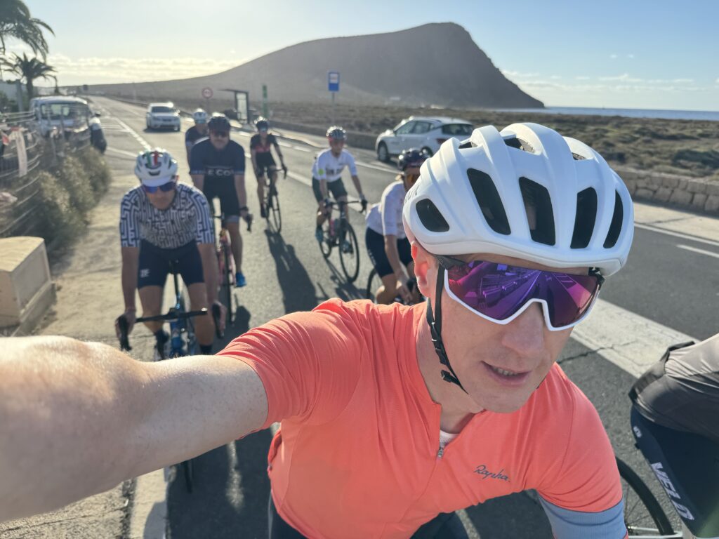 A bunch of cyclists are cruising down a coastal road with a big hill in the background. It's a perfect day—clear blue sky and all. One rider, wearing an orange jersey and white helmet, snaps a selfie from the front.