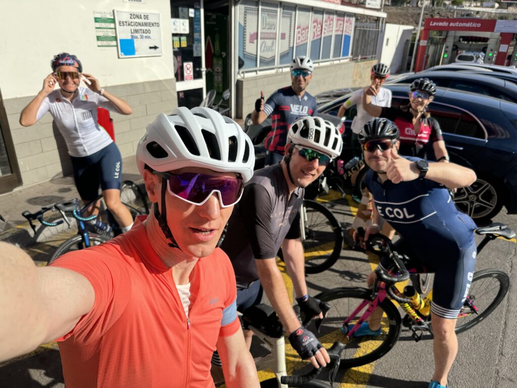 A bunch of seven cyclists in all sorts of colorful gear and helmets are chilling outside a building with their bikes parked nearby, soaking up the sunny vibes and posing for a photo.