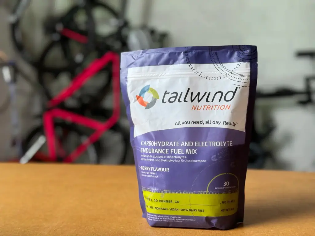 So, there's this pack of Tailwind Nutrition Endurance Fuel Mix in berry flavor chillin' on a wooden table with some blurry bikes in the backdrop. It's basically advertising its carb and electrolyte mix that's perfect for endurance sports.