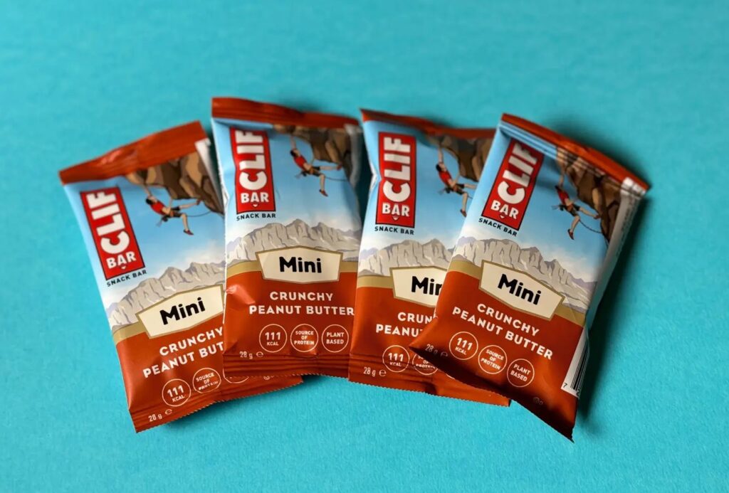 There are four Clif Bar Mini Crunchy Peanut Butter snack bars laid out on a light blue backdrop. The packaging has some cool outdoor adventure pics and shows the nutritional info.