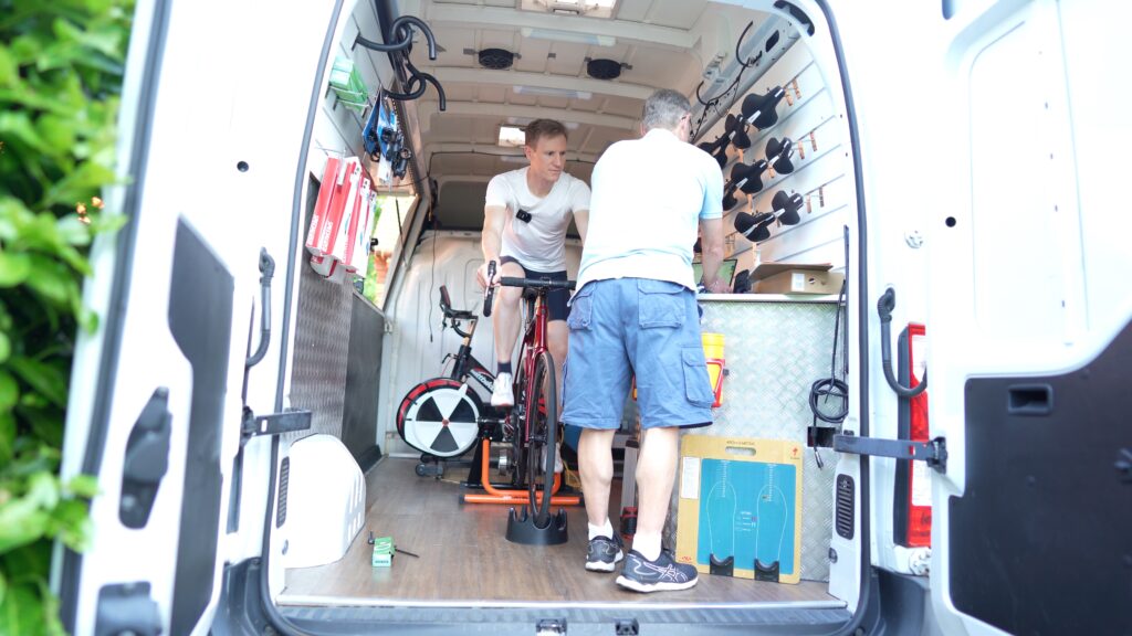 So, there's these two guys in a van that's been tricked out into a traveling bike repair shop. One dude's on a stationary bike, maybe testing or something, and the other guy is helping out with the gear. You can see bikes and tools all over the place.