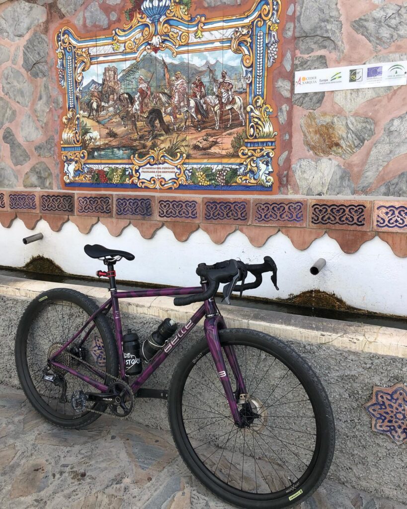 There's a purple bike chilling in front of this cool mural with old-timey folks on it. The mural's stuck to a stone wall, and there's a little fountain trickling underneath.