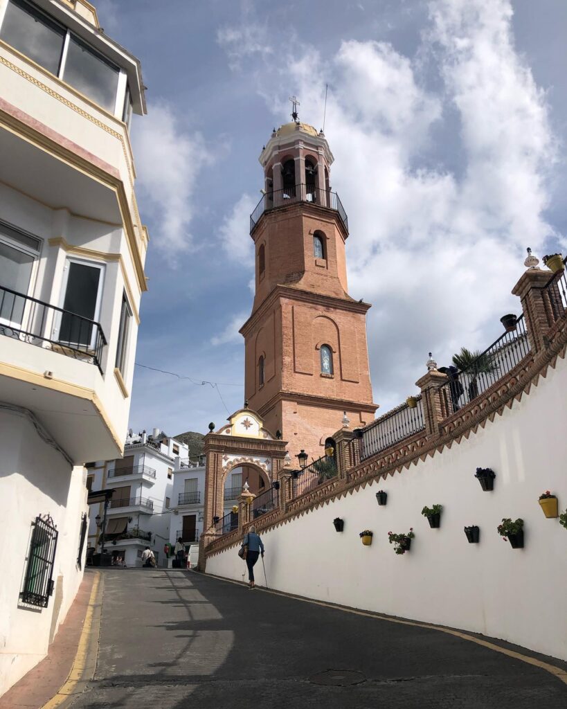 So, someone is strolling up a hilly street, headed towards this big brick bell tower that's got white buildings all around it. And it's a nice sunny day too!