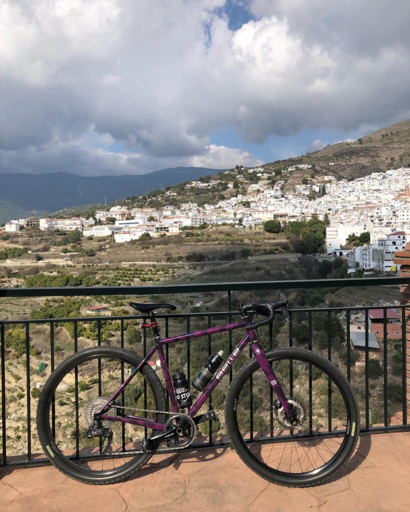 A purple bike is chilling against a railing, overlooking this cool hillside town with white buildings and some random greenery. The sky's partly cloudy, adding to the vibe.