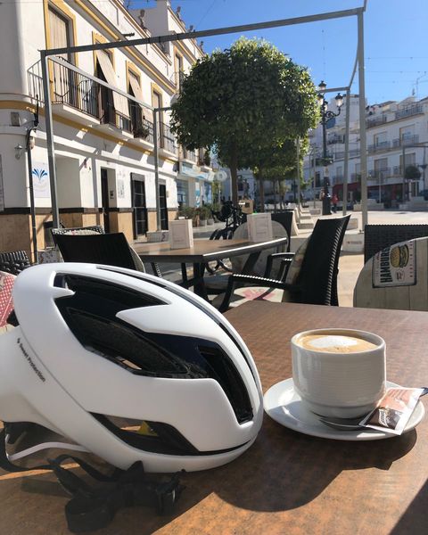 So, there's a white bike helmet chillin' next to a cup of coffee on an outdoor cafe table. In the background, you’ve got a sunny street with some buildings and trees. Just that perfect laid-back vibe!