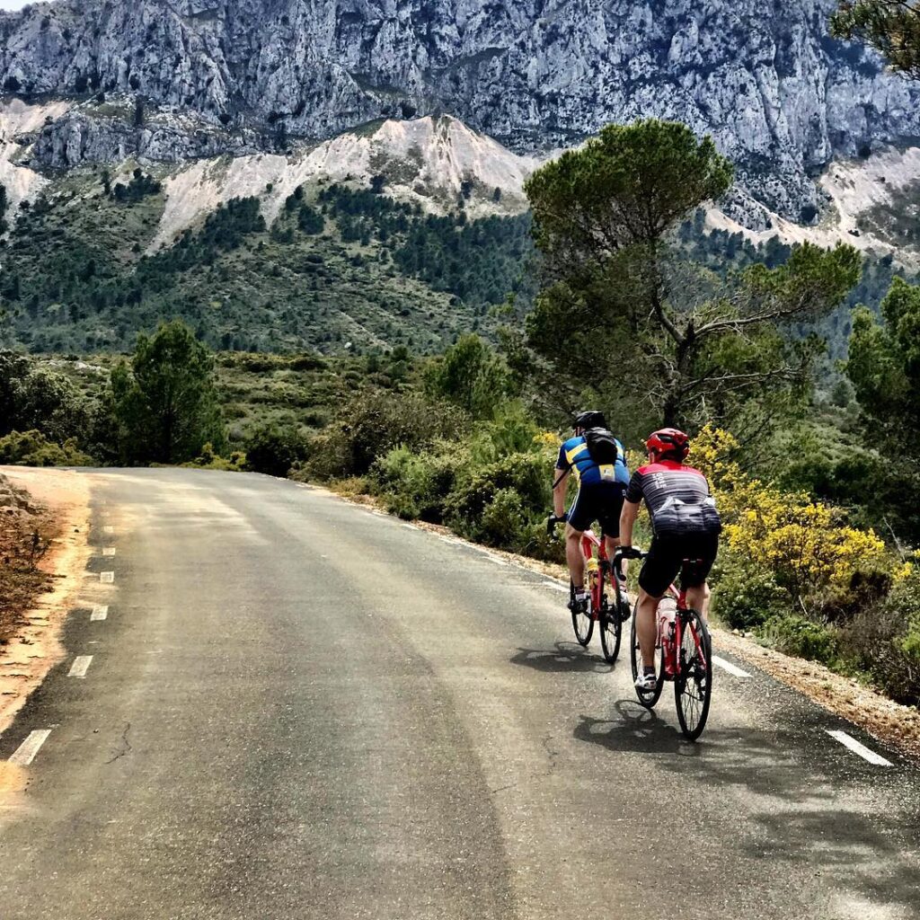 Two cyclists, decked out in their gear, are cruising down a twisty road through some gorgeous mountain scenery, with trees and bushes all around.