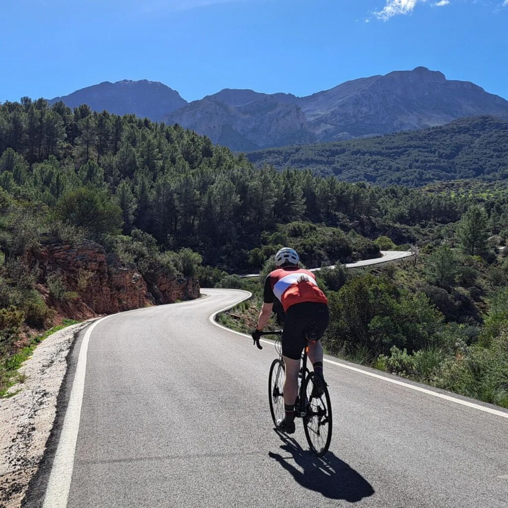 A cyclist is cruising along a twisty mountain road with awesome green scenery all around and a perfect blue sky overhead.