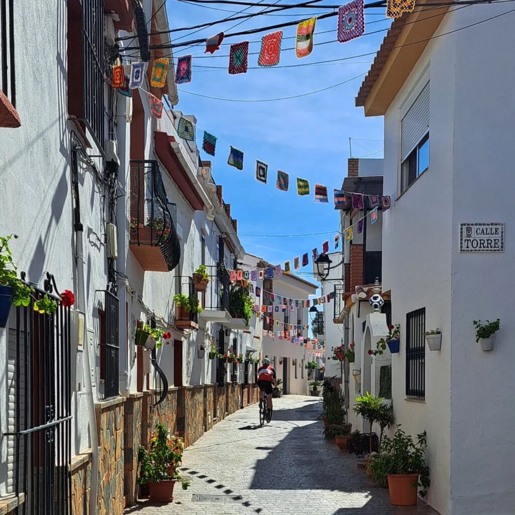 A brightly lit, narrow street with colorful flags strung above and white buildings on either side. A cyclist cruises down the cobblestone path.