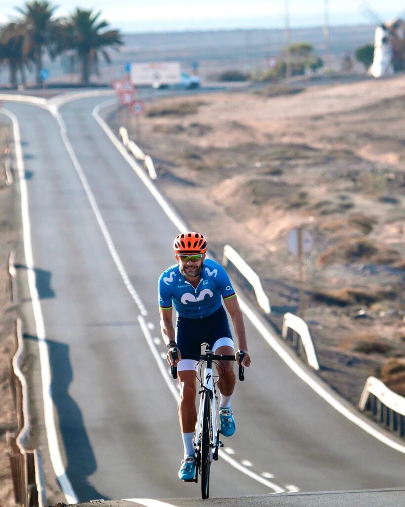 A cyclist in a blue jersey and red helmet is cruising on a quiet, hilly road with palm trees and clear skies around.